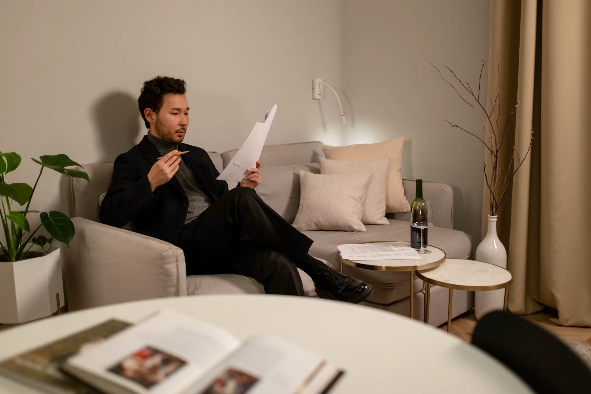 Asian man sitting on a couch reading documents, with a wine bottle on the table.